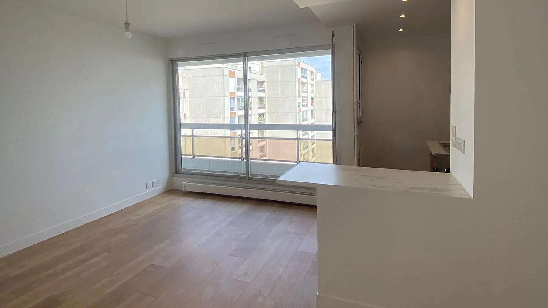 Séjour d'appartement parisien livré après rénovation : murs blancs, parquet clair, baie coulissante sur un balcon et bar en pierre claire ouvert sur la cuisine