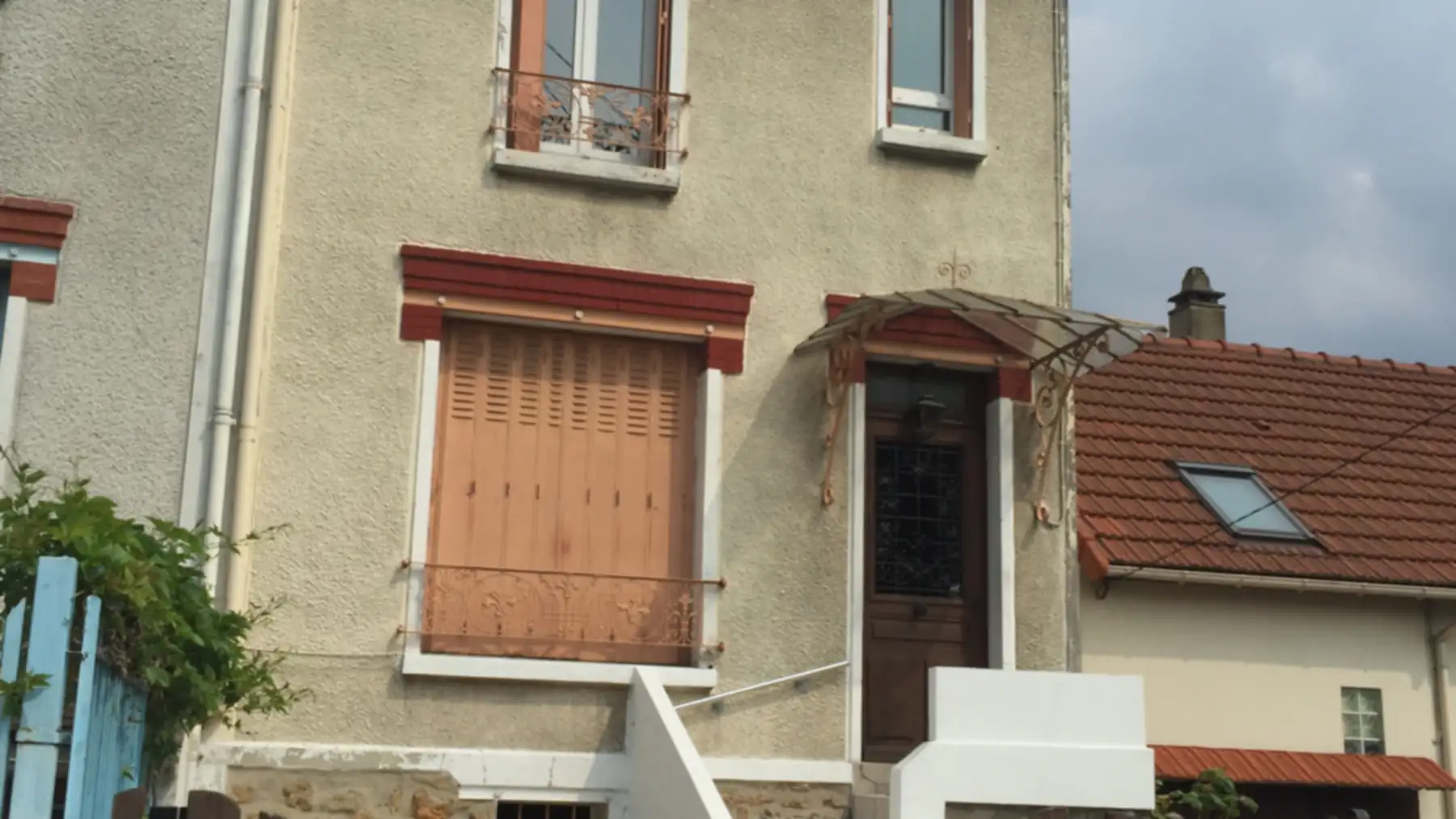 Façade d'une maison de ville de la banlieue parisienne : crépi clair, linteaux de brique rouge, marquise de fer forgé au-dessus d'une porte en bois à vitrail, volet persienne et toit de tuiles mécaniques