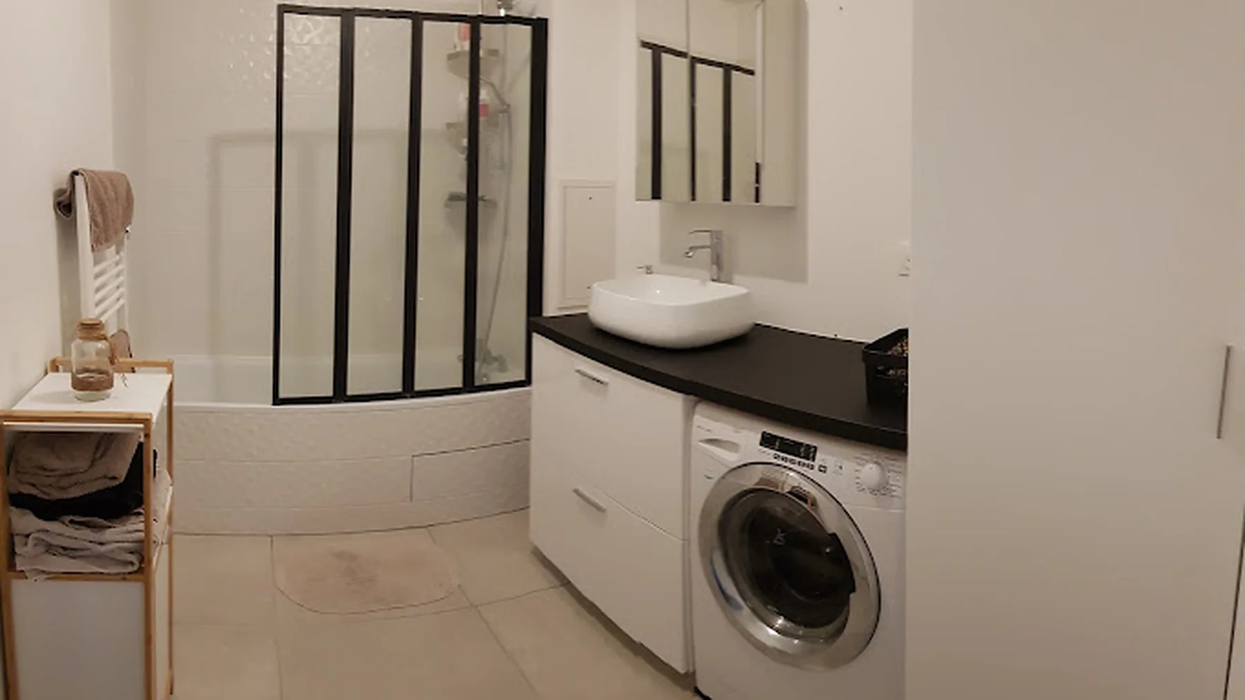 Salle de bain rénovée par Izem Rénovation, pare-baignoire style verrière à cadre noir, faïence blanche à relief, meuble vasque à plan noir et lave-linge encastré sous le plan