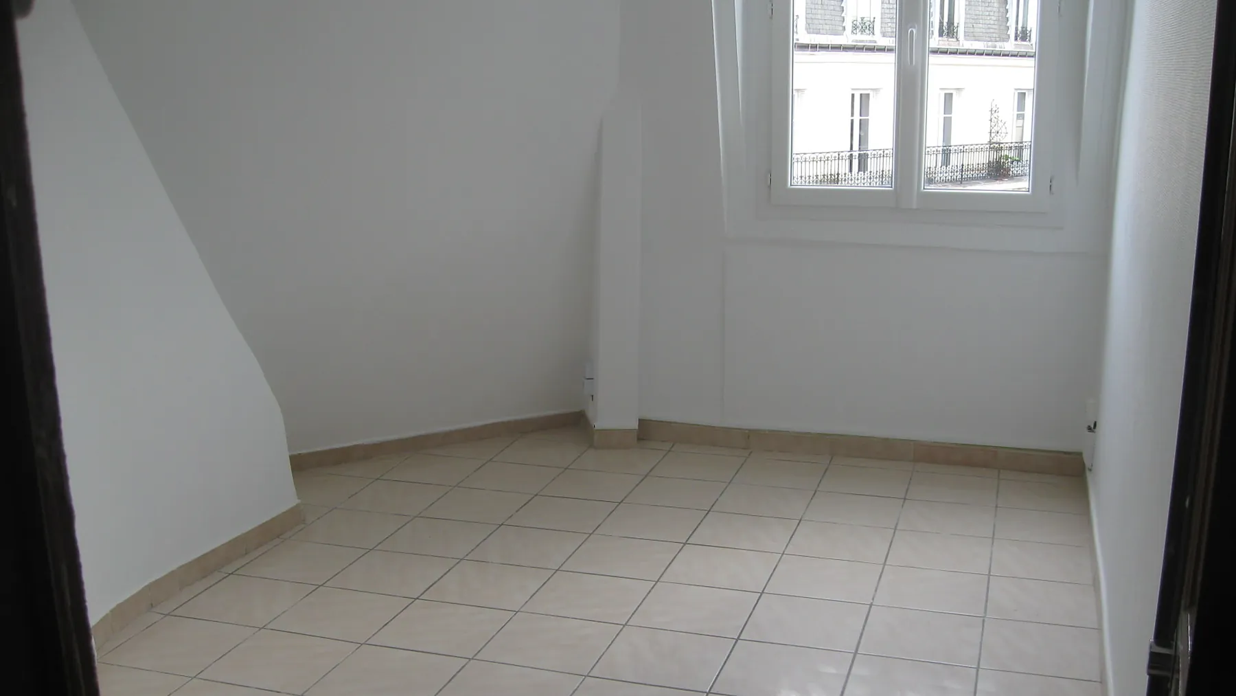 Studio parisien de dernier étage après rénovation, rampant et murs repris en peinture blanche, sol carrelé clair et fenêtre donnant sur les façades d'en face