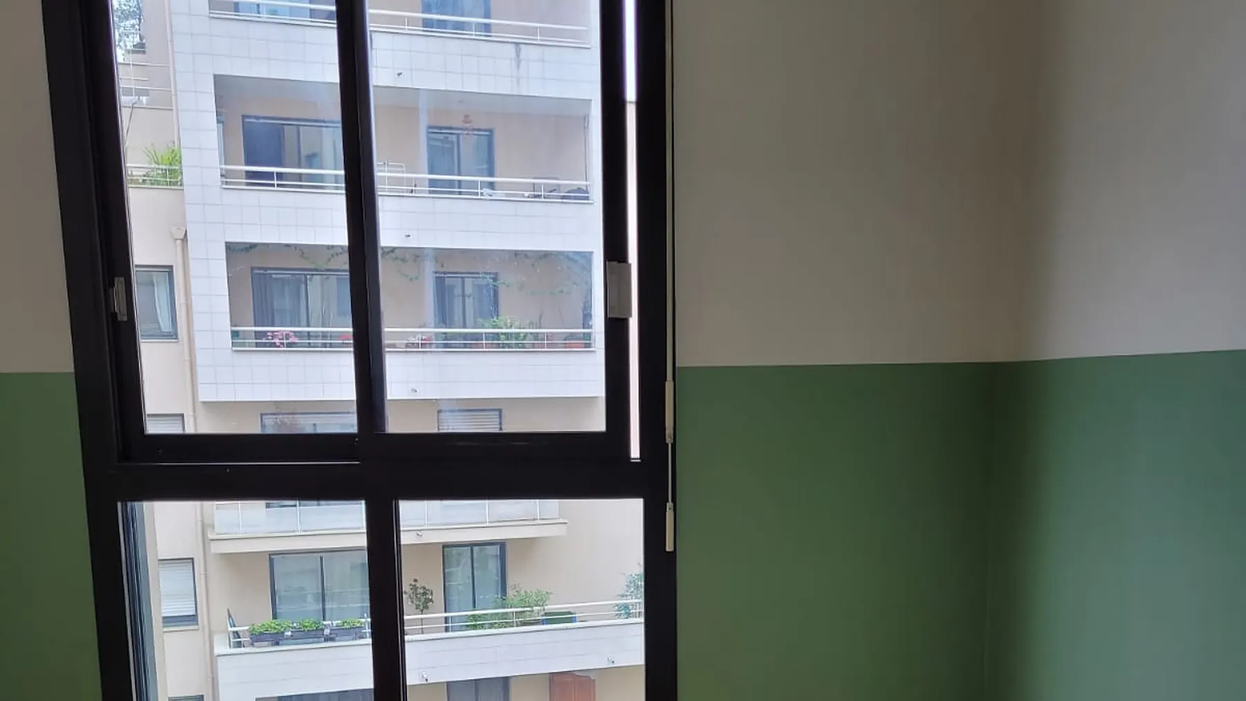 Chambre parisienne repeinte en deux tons, soubassement vert et haut de mur crème, fenêtre à cadre noir ouverte sur la façade à balcons de l'immeuble d'en face
